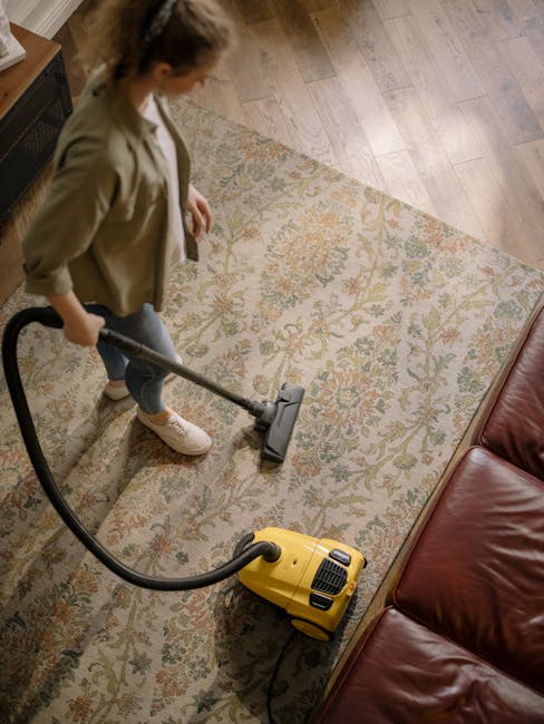 A person with curly hair, wearing a beige jacket and blue jeans, kneeling on a patterned carpet in a room with warm wooden furniture. They are holding the open top of a yellow and black portable vacuu