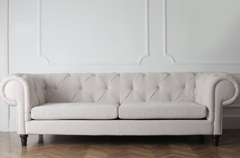 A young boy is using a vacuum cleaner to perform deep surface cleaning and upholstery sanitisation on a beige fabric sofa in a well-lit living room, while an older man lounges on the sofa, appearing r