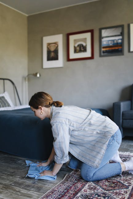 A woman kneeling on a patterned rug in a living room, engaged in surface cleaning by wiping the wooden floor with a blue cloth. She is dressed in a light-colored striped shirt and jeans, with her hair